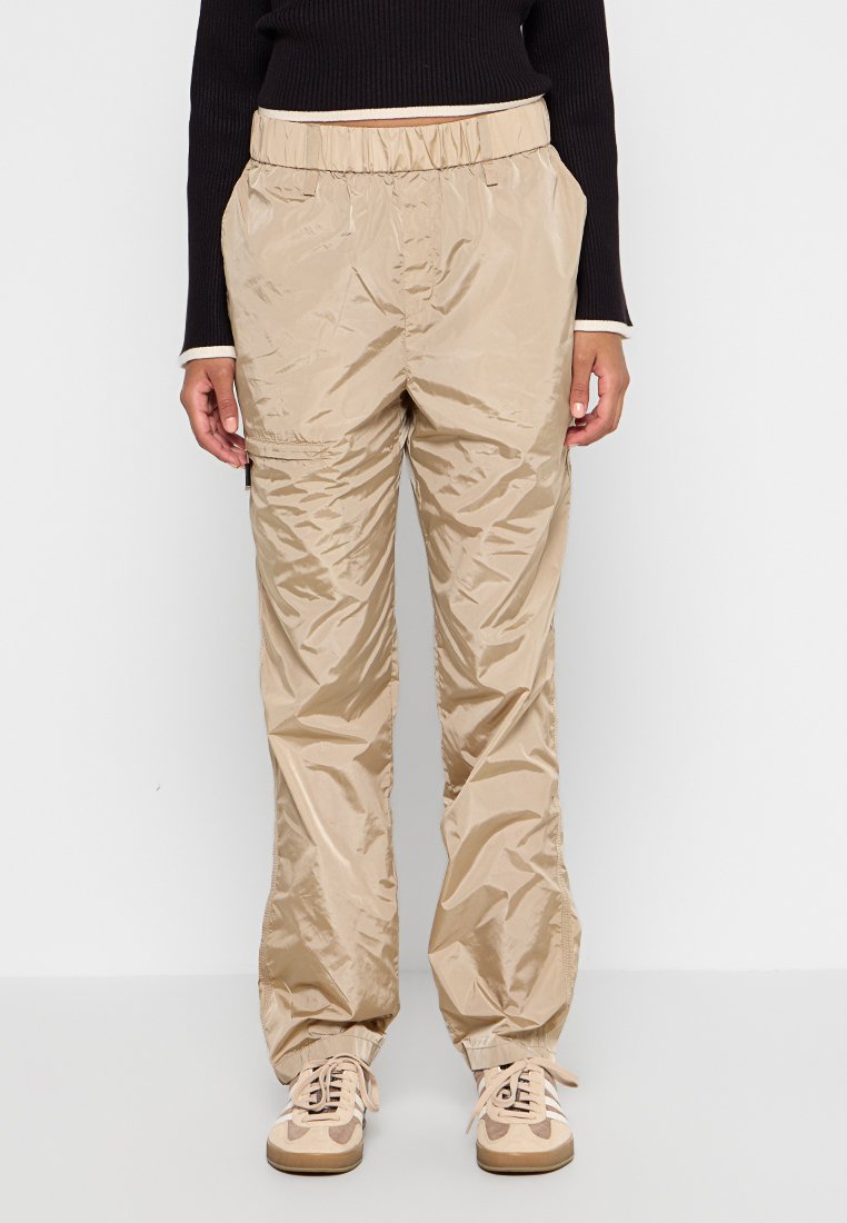 Personne portant un pantalon beige brillant ample avec une ceinture élastique et des baskets beiges, se tenant devant un fond blanc.