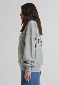 Sudadera gris claro de manga larga, con puños y dobladillo acanalados, que presenta texto oscuro en la parte delantera. Corte casual y holgado.