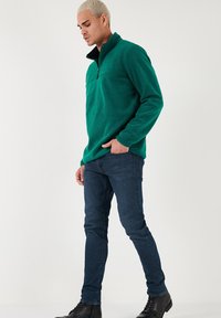 Zeleno flis jopič s zip ovratnikom, ki ima črn akcentski detajl na vratu. Nosi se z modrimi kavbojkami in črnimi usnjenimi škornji.