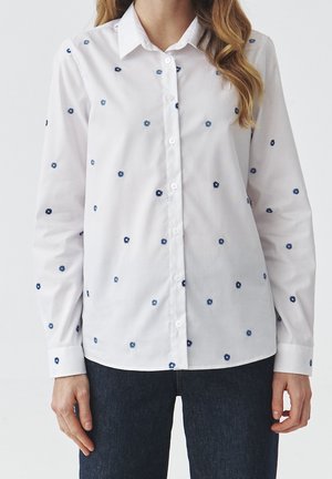Weißes, langärmliges Hemd mit kleinen dunkelblauen Blumenmustern, getragen über dunkelblaue Jeans von einer Person mit hellbraunem, welligem Haar.
