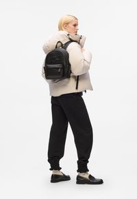 Črni usnjeni nahrbtnik z embosiranim logotipom, sprednja žep z zadrgo, nastavljivi naramnice, nošen čez bež puhovko, črne trenirke in loaferje.