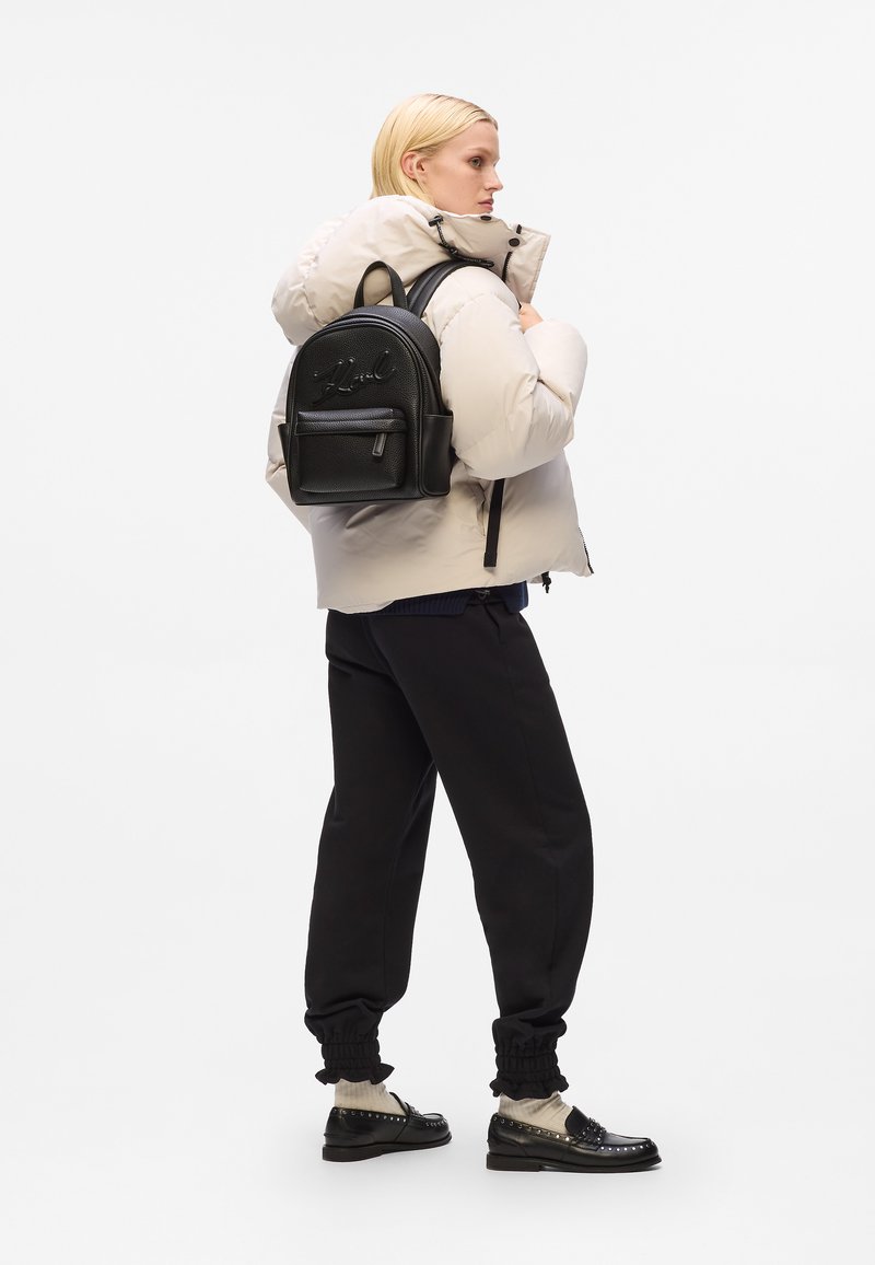 Črni usnjeni nahrbtnik z embosiranim logotipom, sprednja žep z zadrgo, nastavljivi naramnice, nošen čez bež puhovko, črne trenirke in loaferje.