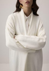 Preveršený biely kardigán z mäkkej, textúrovanej tkaniny, s dlhými rukávmi a voľným strihom, ktorý má rebrované detaily pozdĺž manžet.