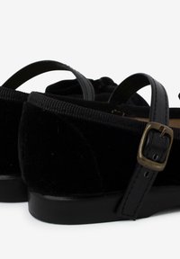 Zapatos de terciopelo negro con punta redondeada, dos correas negras y una hebilla de metal. Presentan una superficie lisa y suela plana.