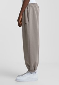 Grå sweatpants med elastisk talje og ribbet manchetter, med sidelommer og et lille logo på låret. Bærer hvide sneakers.