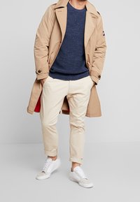 Beige trenchcoat över en marinblå tröja, krämfärgade byxor och vita sneakers. Noterbara detaljer inkluderar en röd foder och uppvikta byxben.