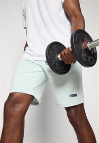 Calções de atletismo verde menta claros com uma cintura elástica e um logótipo branco da Puma. Segurando um peso preto com acabamentos metálicos.