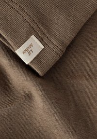 Bruine katoenen stof met een fijne geribbelde textuur, met een label dat in de hoek is genaaid. Het materiaal lijkt zacht en glad.