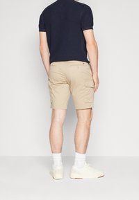 Un homme portant un short cargo beige, des baskets blanches avec une marque visible, des chaussettes blanches montant jusqu'à la cheville, et une chemise courte à manches courtes texturée bleu marine, se tenant de dos.
