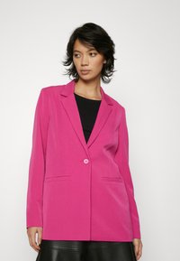 Fitted blazer in bright pink, featuring a single button closure, two front pockets, and a smooth texture. Worn over a black top.