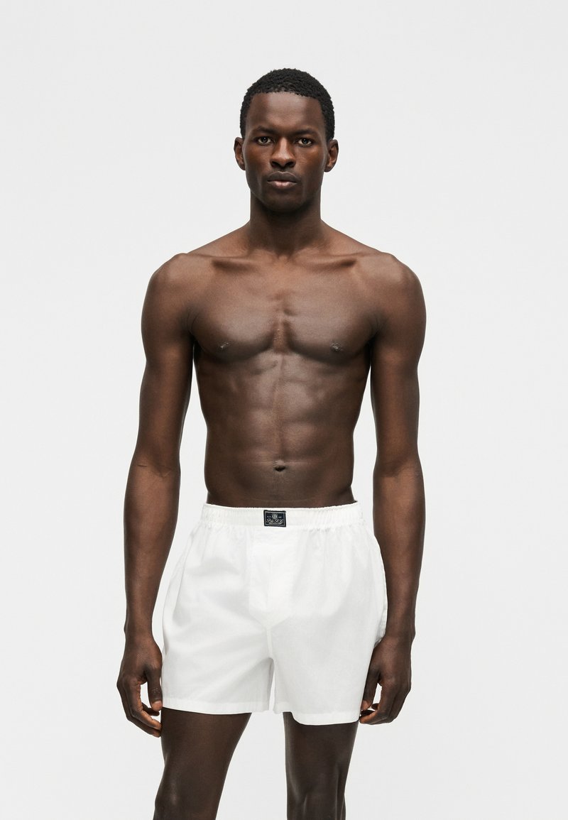 Atletische donkerhuidige man staat tegen een witte achtergrond, gekleed in witte shorts met elastische taille, kijkt recht vooruit met een neutrale uitdrukking en zijn armen langs zijn zij.