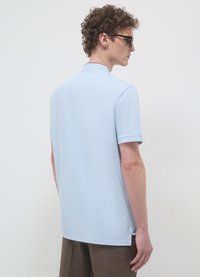 Polo a maniche corte azzurro chiaro con colletto, spacchi laterali sull'orlo e una texture liscia. Il modello indossa occhiali e è di spalle.