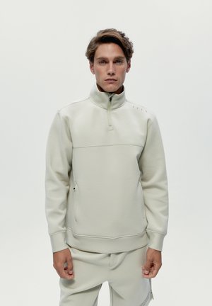 Mann trägt einen hellbeigen Pullover mit Viertel-Reißverschluss und passende Hose, steht vor einem einfarbigen hellen Hintergrund und schaut nach vorne.