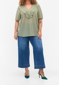 Top verde oliva con pois testurizzati e scollatura a V arricciata, abbinato a jeans blu a gamba larga e décolleté verdi a punta.