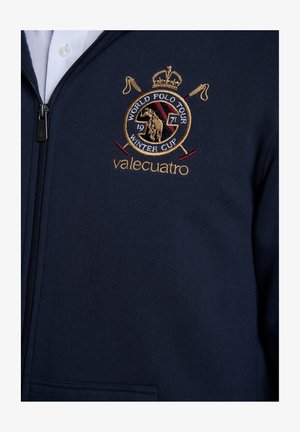 Sudadera con cremallera azul marino hecha de tela suave, con un logo bordado en dorado y burdeos que muestra un caballo y mazos cruzados.