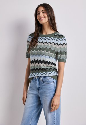Femme souriante aux longs cheveux bruns portant un pull à manches courtes vert, bleu et noir à motif zigzag et un jean bleu clair, debout contre un mur gris.