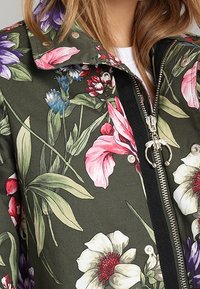 La veste florale présente un tissu vert foncé orné de grandes fleurs roses, violettes et blanches, une fermeture éclair et des accents à boutons-pression.