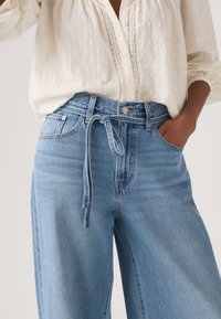 Person wearing a white textured blouse tucked into high-waisted, light blue denim jeans with a tied denim belt and one hand in pocket.