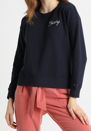 Felpa blu navy con polsini e orlo a costine, con ricamo bianco "Tommy" sul petto, abbinata a pantaloni rosa.