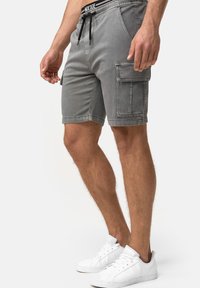 Shorts cargo en denim gris avec des poches latérales, une taille élastique et un cordon de serrage noir. Portés avec des baskets blanches sur un fond neutre.