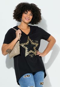 Femme souriante aux cheveux bouclés portant un t-shirt noir à col en V avec un motif d'étoile dorée, un jean déchiré, et tenant un sac pailleté doré sur l'épaule.