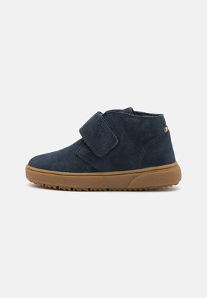 Chaussure cheville en daim bleu marine avec une large bande velcro et une semelle épaisse en caoutchouc marron, vue de côté sur un fond uni.