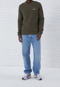 Suéter de canalé verde oliva con cuello redondo, combinado con jeans de pierna recta azul claro y zapatillas de running metálicas sobre un fondo de ladrillo blanco.