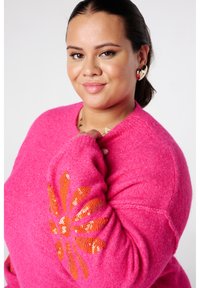 Lachende vrouw met donker haar in een staart draagt gouden oorbellen in de vorm van een hart en een felroze trui met een oranje paillettenbloem op de mouw.