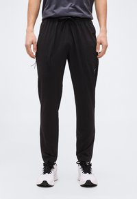 Άνδρας που φοράει μαύρο αθλητικό παντελόνι με κορδόνι και λευκά παπούτσια τρεξίματος, στέκεται σε απλό ανοιχτό φόντο.