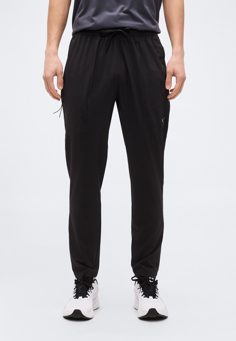 Άνδρας που φοράει μαύρο αθλητικό παντελόνι με κορδόνι και λευκά παπούτσια τρεξίματος, στέκεται σε απλό ανοιχτό φόντο.