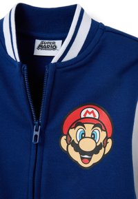 Chaqueta azul con cremallera, cuello a rayas blancas y una impresión de la cara de Mario con un sombrero rojo que tiene el emblema "M" en el pecho.