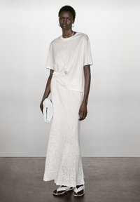 Hellgraues, oversized T-Shirt, kombiniert mit einem langen, weißen, strukturierten Häkelrock, während sie eine weiße Perlen-Clutch hält und flache Sandalen trägt.
