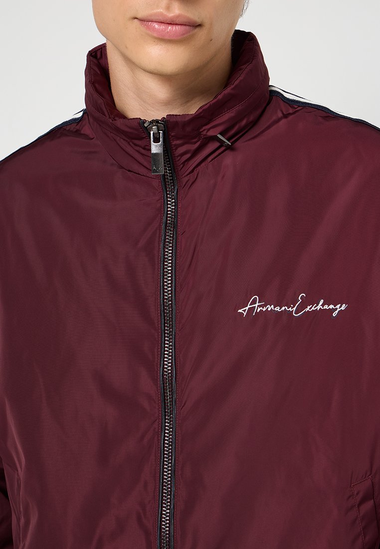 Giacca bomber bordeaux con colletto alto, chiusura a zip e logo ricamato in bianco. Presenta un tessuto leggero e lucido con strisce in contrasto.