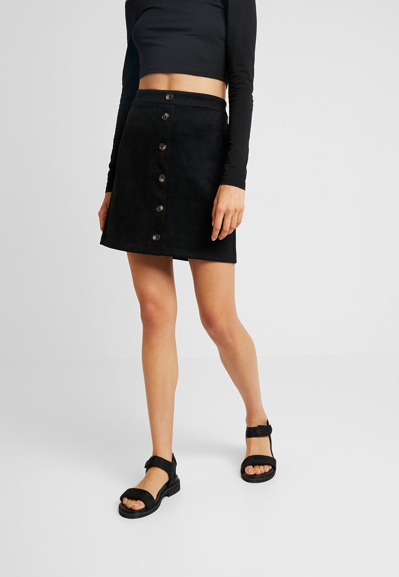 Femme portant un top noir à manches longues cropped, une mini-jupe noire à boutons sur le devant et des sandales noires, se tenant sur un fond uni.