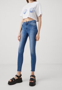 Camiseta corta blanca con bordado de concha azul, combinada con jeans ajustados azules con dobladillo deshilachado y sandalias negras gruesas.