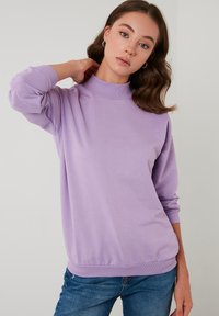Felpa lilla con collo alto e polsini a costine. Tessuto morbido con una vestibilità comoda e orlo a banda. Indossata con jeans blu.