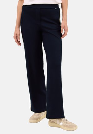Vrouw in een marineblauwe wijde broek met witte zijkanten en beige sneakers, staand tegen een lichte achtergrond.