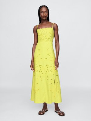 Femme avec des cheveux tressés portant une robe maxi sans manches de couleur vert lime avec des motifs floraux découpés et des sandales à brides transparentes, se tenant sur un fond gris.