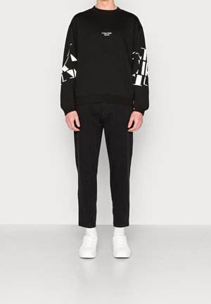 Schwarzes Sweatshirt mit weißem Grafikdruck auf den Ärmeln, Rundhalskragen, kombiniert mit schwarzen Hosen und weißen Sneakers. Einfaches und modernes Design.