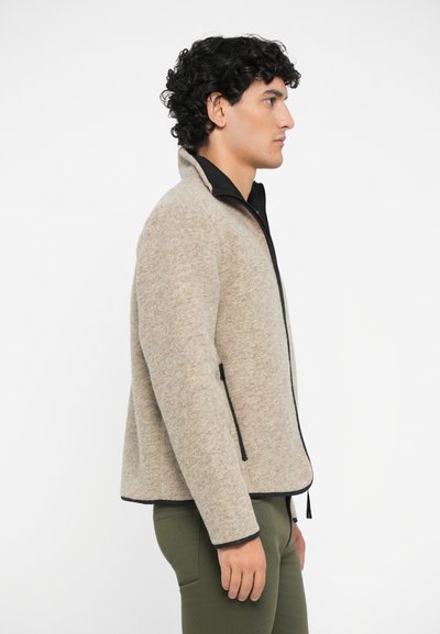 Beige fleece jacket with a black trim and zippered pockets, featuring a high collar and a structured fit. Material appears soft and textured.