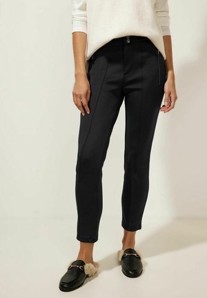 Pantalones negros ajustados con un corte slim, textura suave, bolsillos frontales y cierre con botón, combinados con mocasines oscuros.