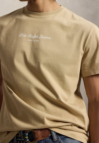 Άνδρας που φοράει μπεζ μπλουζάκι Polo Ralph Lauren, μέσα σε μπλε τζιν με δερμάτινη ζώνη με αγκράφα και ριγωτές λεπτομέρειες.