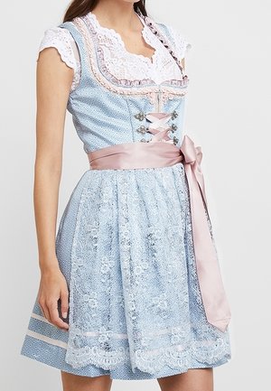 Kvinde iført en blå mønstret dirndl-kjole med hvid blonderbluse og lyserødt satinbånd bundet om taljen.