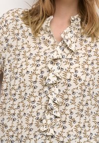 Kwiatowa bluzka z lekkiej kremowej tkaniny, z falbanką przy zapięciu, brązowymi i niebieskimi wzorami kwiatowymi oraz kołnierzykiem mandarynkowym.