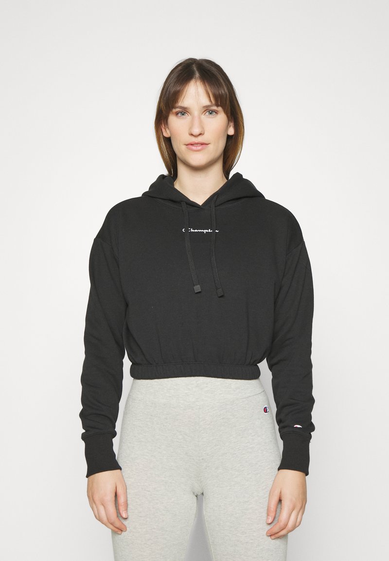 Felpa corta nera in cotone, con cappuccio regolabile e logo ricamato sul davanti, abbinata a leggings grigi.