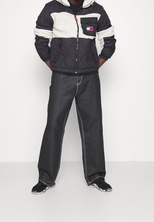 Mann trägt eine schwarz-weiße gesteppte Jacke mit einem Tommy Jeans Logo, weite schwarze Hosen mit weißen Nähten und schwarze Sneakers mit gemusterten Sohlen.