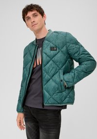 Grüne gesteppte Jacke mit Reißverschluss, geripptem Kragen und Vordertaschen. Sie hat eine strukturierte Oberfläche und wird mit einem dunklen T-Shirt kombiniert.