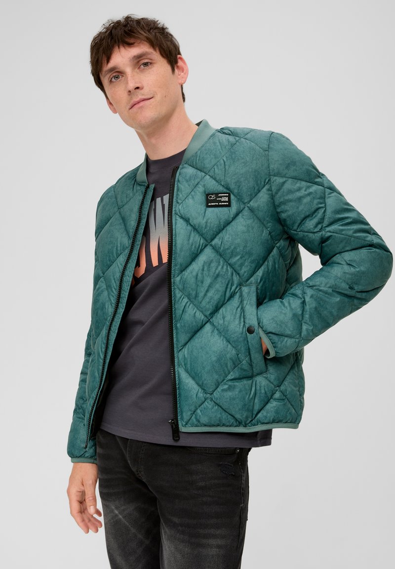 Grüne gesteppte Jacke mit Reißverschluss, geripptem Kragen und Vordertaschen. Sie hat eine strukturierte Oberfläche und wird mit einem dunklen T-Shirt kombiniert.