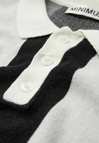 Polo noir et crème, avec un tissu texturé et trois boutons transparents à la patte de boutonnage ainsi qu'un col. Étiquette de marque visible.