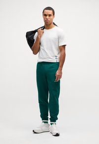 Zöld melegítőnadrág rugalmas mandzsettával és Calvin Klein logóval. Fehér kerek nyakú póló. Fekete táska vállon átvetve. Fehér tornacipő.